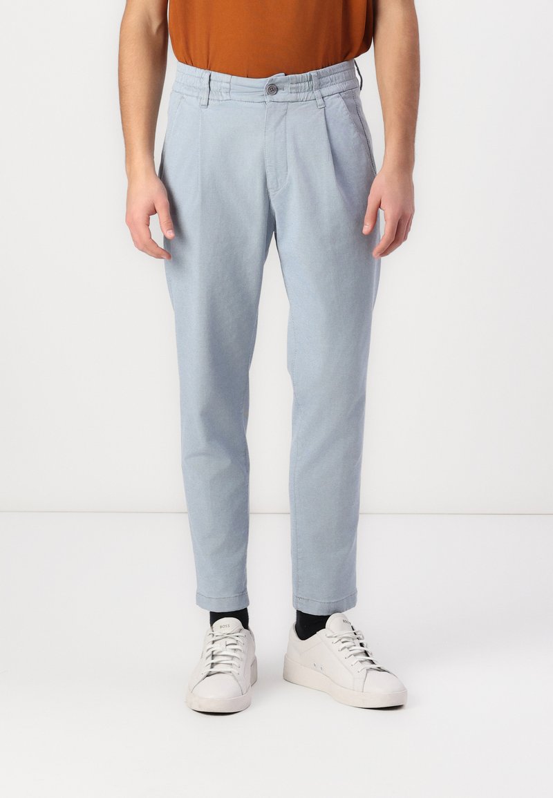 Pantalons ajustés bleu clair avec plis, en tissu lisse, dotés d'une fermeture à bouton et de chevilles fuselées, associés à des baskets blanches.