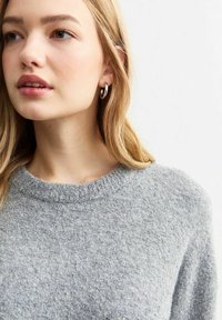 Grå stickad tröja med rund halsringning, mjuk textur och ribbad detalj runt kragen. Silverfärgade hoops örhängen bärs.