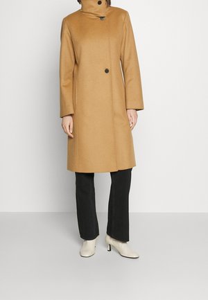 Frau trägt einen langen Kamelmantel mit hohem Kragen, schwarze Hose und weiße Stiefeletten mit Absatz, steht vor einem schlichten Hintergrund.