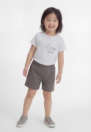 Niño sonriente de pie con las manos relajadas, vistiendo una camiseta gris clara con caras de animales y "Somos familia", pantalones cortos grises y zapatos sin cordones.