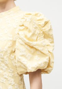 Κίτρινο φόρεμα με υφή από λουλουδάτα σχέδια και μεγάλο φουσκωτό μανίκι που καλύπτει το πάνω μέρος του βραχίονα και τον ώμο.