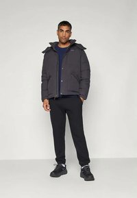 Ενισχυμένο μπουφάν σκούρου γκρι χρώματος με κουκούλα, μπροστινή κλείσιμο με κουμπιά, φορεμένο πάνω από μπλε πουκάμισο, συνδυασμένο με μαύρο παντελόνι και μαύρα αθλητικά παπούτσια.