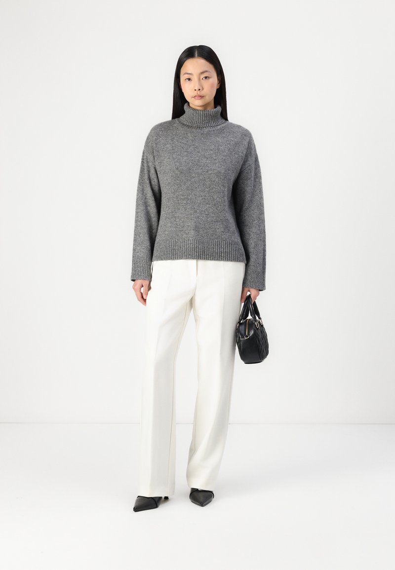 Frau trägt einen grauen Rollkragenpullover, weiße weitgeschnittene Hose, schwarze spitze Schuhe und hält eine schwarze Handtasche vor einem weißen Hintergrund.