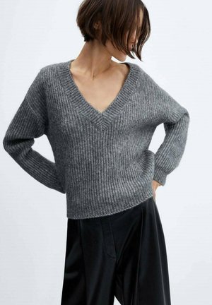 Woman with short hair wearing a grey ribbed V-neck sweater and black pants, posing with hands on hips against a white background.