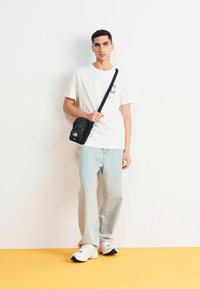 Borsa a tracolla nera, t-shirt bianca con logo, jeans azzurri chiari e sneakers bianche. Completo semplice e casual su uno sfondo neutro.