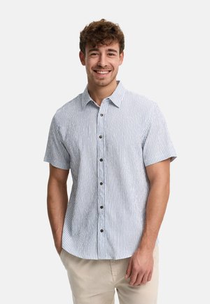 Homme souriant aux cheveux bouclés portant une chemise à manches courtes boutonnée à rayures bleu clair et blanc, et un pantalon beige, debout les mains dans les poches.