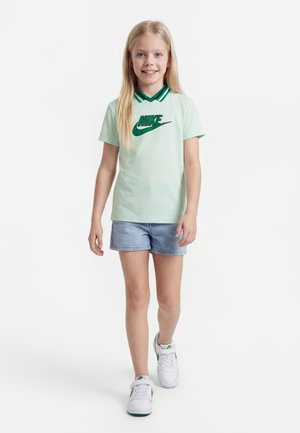 Jeune fille aux longs cheveux blonds portant un t-shirt Nike vert clair, un short en denim et des baskets blanches, marchant vers l'avant sur un fond blanc.