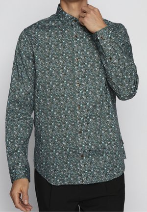 Mężczyzna poprawiający kołnierz koszuli na guziki z długim rękawem, ozdobionej ciemnozielonym i niebieskim kwiatowym wzorem, noszący czarne spodnie na jednolitym tle.