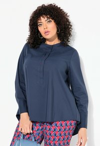 Marineblaue Langarmbluse mit hohem Hals und Frontknopfverschluss, verziert mit roten Akzenten. Kombiniert mit gemusterten roten und blauen Hosen.