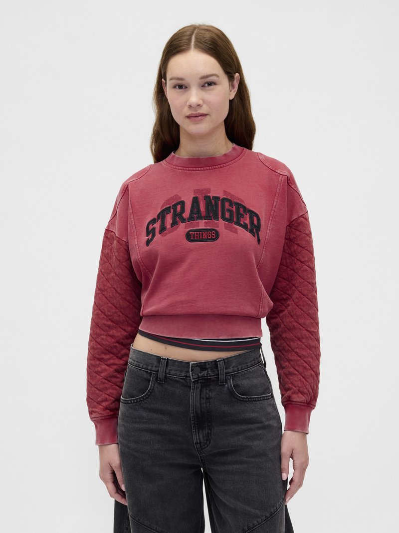 Sudadera corta roja con texto negro "STRANGER THINGS"; mangas acolchadas; textura suave; combinada con jeans negros de talle alto.