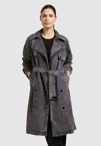 Donna che indossa un lungo trench in denim nero sbiadito con cintura, tasche e una camicia nera sotto, in piedi davanti a uno sfondo semplice.