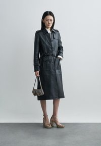 Black leather trench coat with a belted waist, double-breasted buttons, and side pockets. The model holds a small gray handbag.