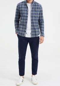 Košile s knoflíky a károvaným vzorem v modrých a zelených tónech, pod ní bílý tričko, doplněno tmavě modrými kalhotami a bílými teniskami. Hladká bavlněná textura.