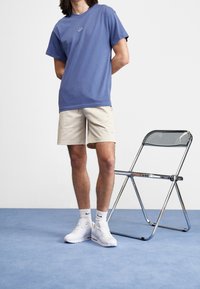 Camiseta de manga corta morada con el logo de Nike en blanco, pantalones cortos beiges, zapatillas blancas y calcetines blancos, de pie junto a una silla plegable de cromo.