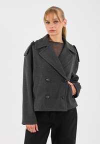 Una donna con capelli biondi indossa un cappotto di lana grigio scuro doppiopetto e pantaloni neri, in piedi davanti a uno sfondo bianco uniforme.