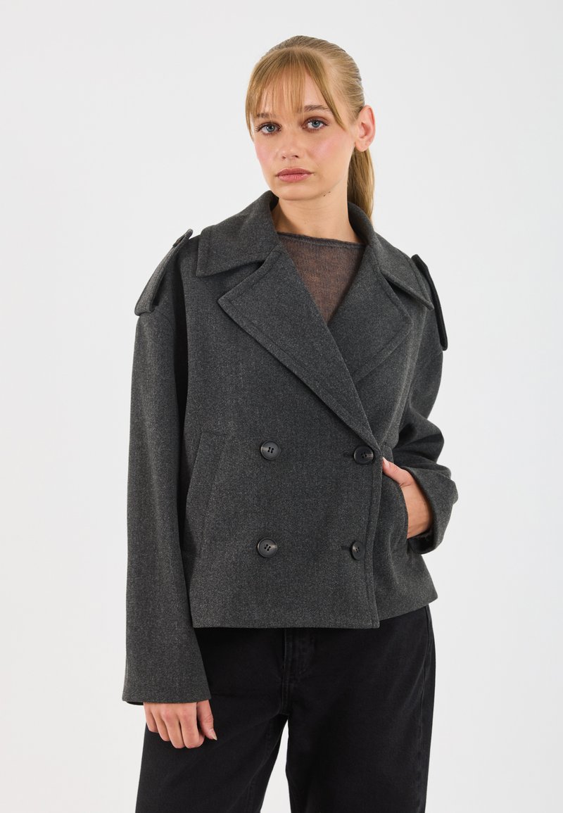 Una donna con capelli biondi indossa un cappotto di lana grigio scuro doppiopetto e pantaloni neri, in piedi davanti a uno sfondo bianco uniforme.