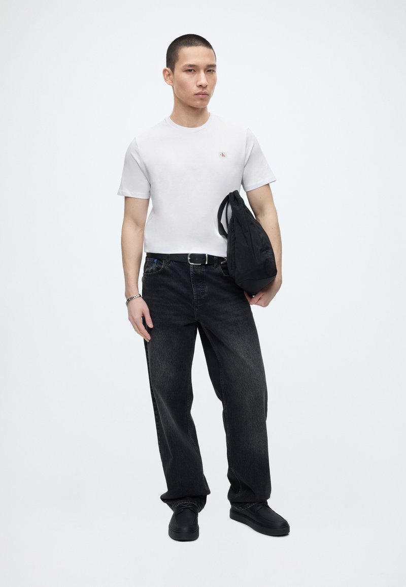 Homme portant une chemise blanche à manches courtes, ceinture noire, jean noir délavé, chaussures noires, tenant un sac noir sur la hanche, debout devant un fond blanc uni.