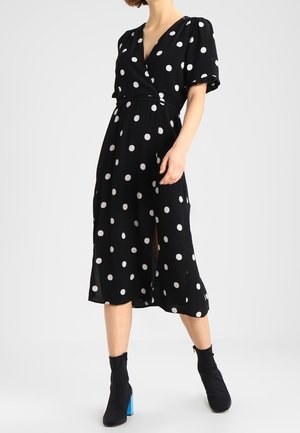 Femme portant une robe midi noire à pois blancs, manches courtes bouffantes, fente devant et bottines noires à talons bleus.
