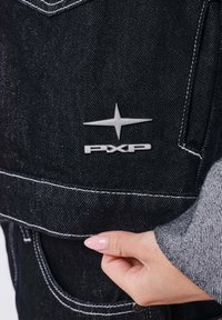 Main tenant un tissu en denim noir avec des coutures blanches et un logo PXP argenté avec un emblème étoile sur le vêtement.