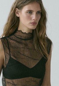 Černý průsvitný top s vysokým límcem a vroubkovaným lemem, zdobený texturovaným vzorem a vrstveným designem přes černou podprsenku ve stejné barvě.