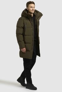 Cappotto puffer verde oliva con cappuccio, zip frontale e bottoni a pressione. Presenta sezioni trapuntate e tasche laterali. Indossato sopra un abbigliamento nero.