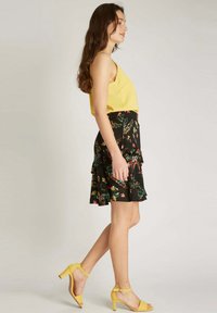 Haut sans manches jaune associé à une jupe noire à fleurs avec des volants empilés ; le mannequin porte des sandales à talons jaunes.