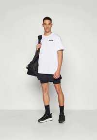 Biela tričko s krátkym rukávom a čiernym logom, v kombinácii s tmavými šortkami a čiernymi teniskami. Model drží čiernu športovú tašku.