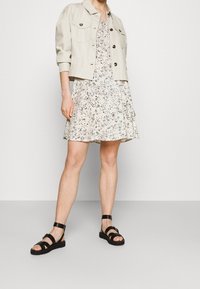 Jasno-beżowa kurtka zapinana na guziki z dwoma kieszeniami na piersi, w połączeniu z sukienką w kwiatowy wzór w kolorze kremowym oraz czarnymi sandałami na płaskiej podeszwie.