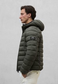 Grüne Pufferjacke mit Kapuze, mit gesteppten Abschnitten, einem Reißverschluss und einem Logopatch am linken Ärmel. Glatte Textur und leicht.