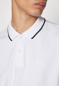 Vit pikétröja med texturerad tyg, svart kant på kragen och tvåknäppning. Har en präglad logotyp på bröstet.