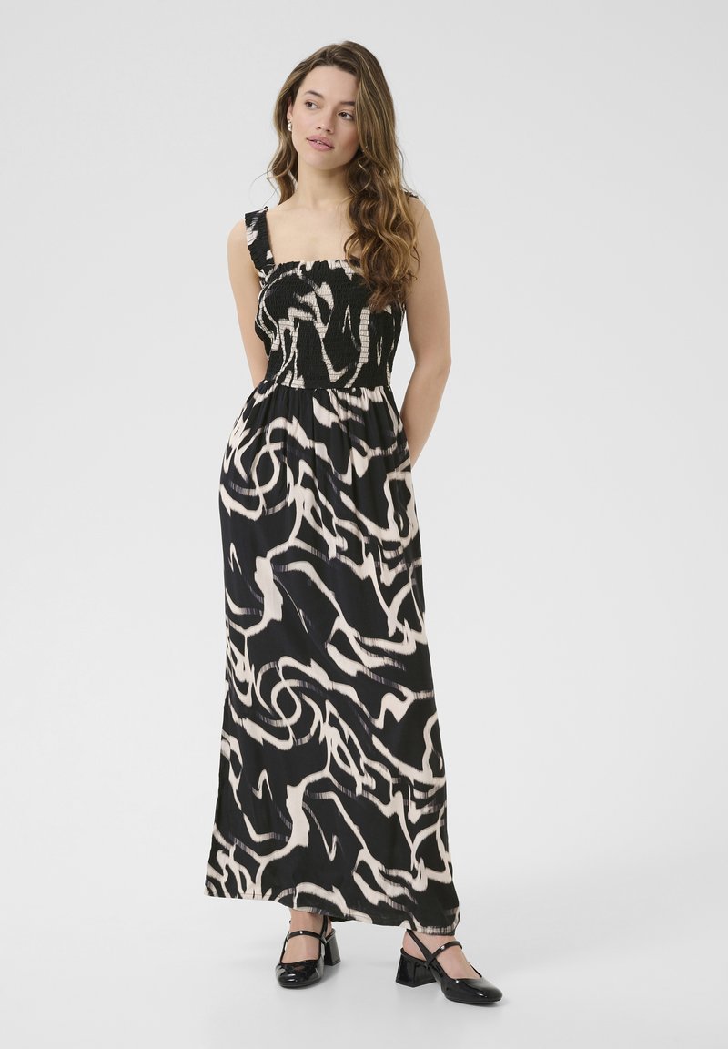 Robe maxi avec un motif abstrait noir et blanc, un corsage smocké, de larges bretelles et une silhouette fluide. Associée à des chaussures plates noires.
