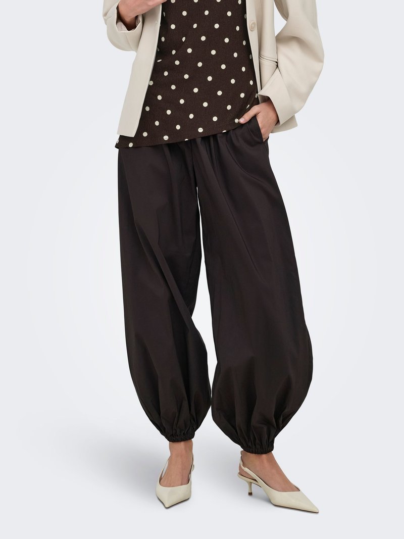 Femme portant un blazer beige, un top marron à pois, un pantalon noir ample plissé et des talons slingback blancs pointus, debout avec une main dans la poche.