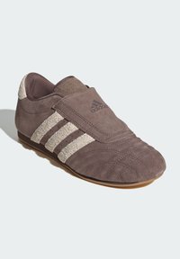 Brown suede shoe with a textured finish, featuring three light beige stripes, a round toe, and a short, flat sole. Brand logo on upper.