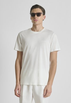 Uomo che indossa una maglietta beige chiaro e pantaloni, con occhiali da sole scuri, in piedi contro uno sfondo grigio uniforme, rivolto in avanti.