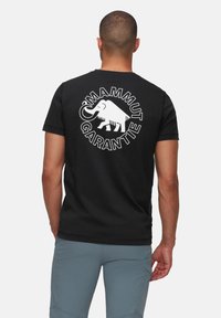 Zwarte katoenen t-shirt met een grote witte afbeelding van een mammoet en de tekst "Mammut Garantie" op de achterkant, korte mouwen en een ronde hals.