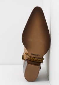 Zapato de tacón alto marrón con parte superior de cuero amarillo texturizado. Presenta una puntera curvada y puntiaguda, además de una correa con detalle de hebilla, etiquetado "BRONX" en la suela.