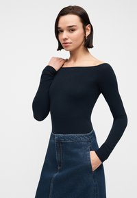 Νεαρή γυναίκα με κοντά σκοτεινά μαλλιά, φορώντας εφαρμοστή μαύρη μπλούζα με μακριά μανίκια και σκέρτσο ψηλόμεσο από σκούρο τζιν, ποζάροντας με το ένα χέρι στον ώμο.