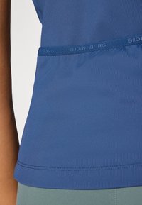 Mėlynas gurguliuojantis tank topas, pasižymintis tekstūruota medžiaga, BJORNBORG logotipo juosta ir prigludusia forma su įlinkusia apatine briauna.