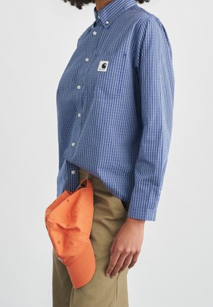 Person iført en blåstribet skjorte med knapper og kaki bukser, der holder en orange baseballkasket ved siden af sig mod en ensfarvet baggrund.