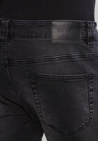 Bakvy av svarta denimjeans med en lädermärke som har texten "ONLY & SONS" ovanför den högra bakfickan.
