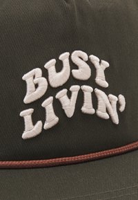 Mørkegrønn caps laget av stoff med hvit brodert tekst "BUSY LIVIN'," og et brunt vevd akcentbånd langs bunnen.