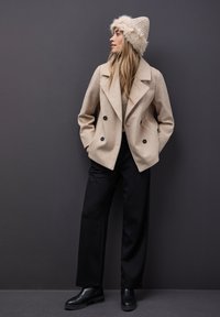 Beige dvojřadový kabát z vlněné směsi s černými knoflíky, černé široké kalhoty a světle huňatou pletenou čepici. Černé kotníkové boty dokončují outfit.