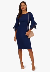 Navyblaues Kleid mit ausgestellten, durchsichtigen Ärmeln, taillierter Schnitt, knielang, kombiniert mit einer beigen Clutch und nude-farbenen, trägerlosen High Heels.