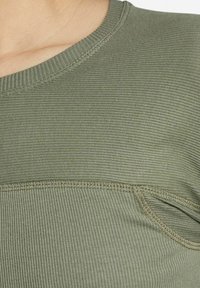Olivgrön ribbad stickad topp med rund halsringning, med subtil textur och kontrasterande sömnadsdetaljer längs sömmarna.