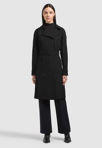 Donna che indossa un trench nero con cintura e bottoni doppiopetto, pantaloni neri a gamba larga e stivali neri, in piedi davanti a uno sfondo semplice.