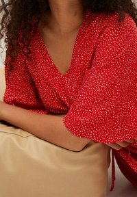 Haut rouge en v avec un enroulement et des manches courtes, présentant un motif à pois blancs. Les bras du mannequin reposent sur un sac beige lisse.