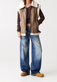 Gilet marrone senza maniche con colletto in shearling sopra un maglione scuro, abbinato a jeans a gamba larga blu sbiaditi e stivali beige.