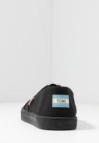 Svart kanvassko med en strukturerad yta, rund form, platt gummisåle och en ljusblå etikett som står "TOMS" på hälen.