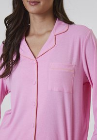 Rosa pyjamasöverdel med ribbad textur, notch-krage med orange piping, en framficka och knappstängning.