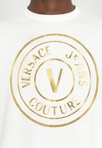 Balts kokvilnas T-krekls arapaļo zelta folijas logotipu, uz kura rakstīts "VERSACE JEANS COUTURE" un centrā ir bolds "V".